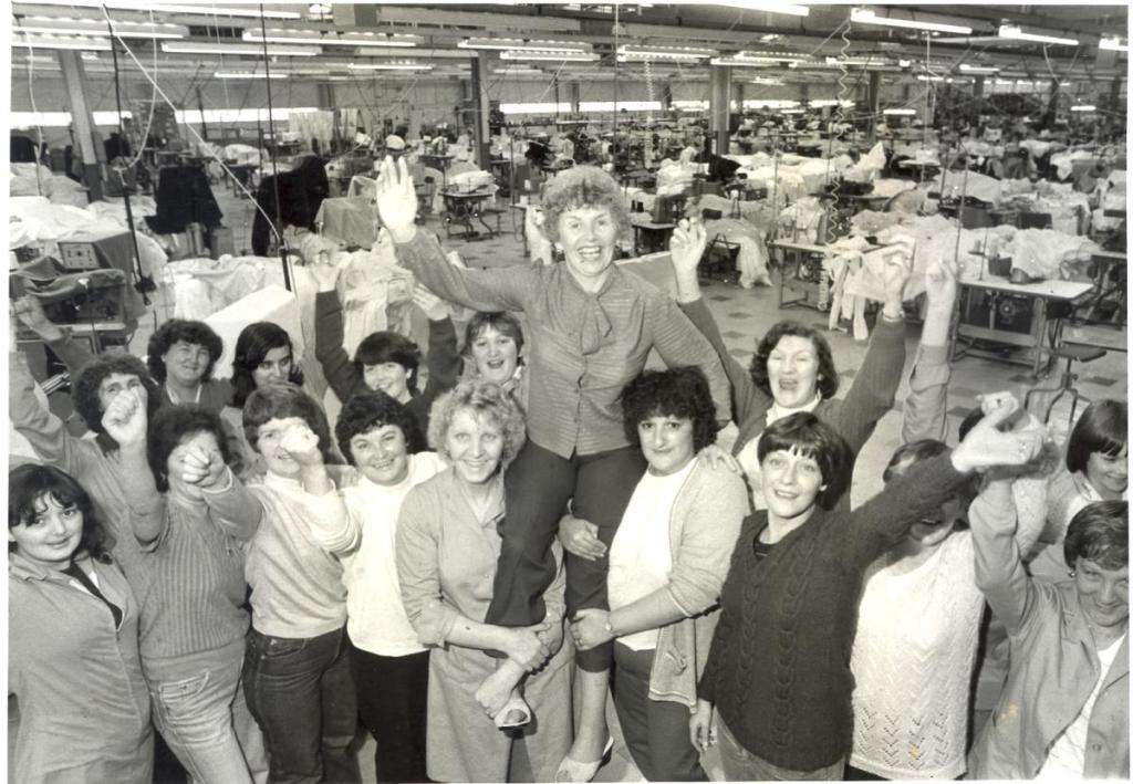A group of 16 women hoist another woman on their shoulders. All are smiling and raising a hand either to wave or as a fist in celebration. The Lee Jeans factory floor with machines can be seen behind them.