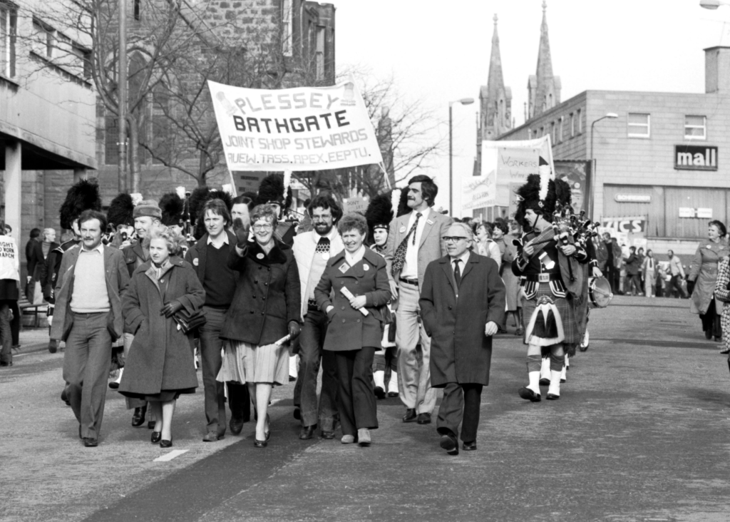 Workers from Plessey Bathgate march with their union banner towards the camera. The picture is in black and white. They are accompanied by pipers in kilts and highland regalia.