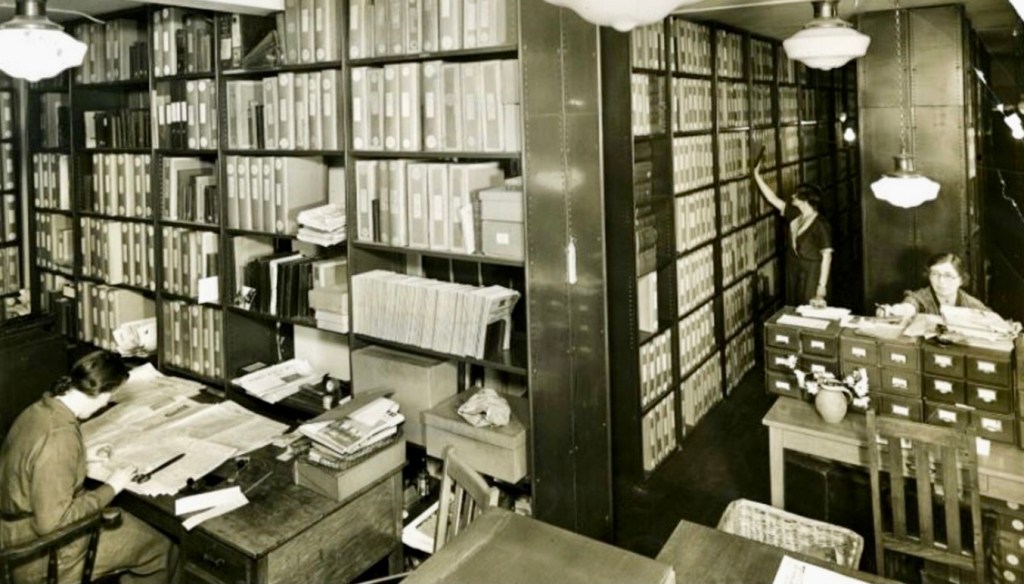 Black and white photo showing two women working in an old fashioned academic library, with shelves from floor to ceiling holding folders. One one table is a set of drawers for index cards.