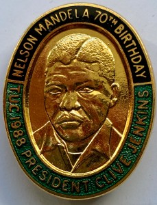 The Mandela badge is a large uppity oval shape. The outer band is in two halves: the top half is black enamel with the words ‘Nelson Mandela 70th birthday’; the lower half is green enamel with the words ‘TUC 1988 President Clive Jenkins’. A central image shows a head and shoulders portrait of Mandela in raised gold set against a gold background.