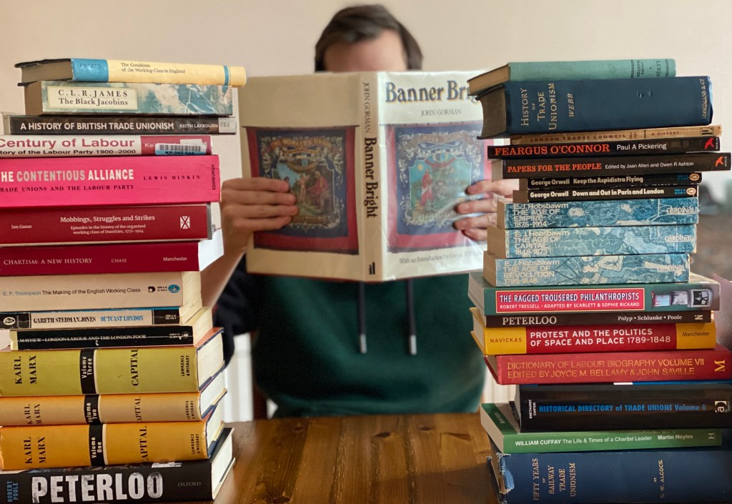 Young man sits facing the camera, his face largely obscured by a copy of the book Banner Bright. To his right and left are teetering piles of labour history books.