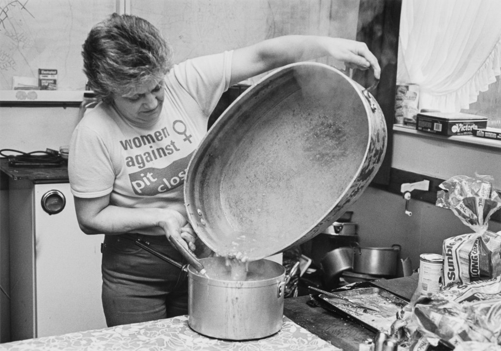 A woman wearing a Women Against Pit Closures t-shirt pours food from a huge bowl into a saucepan.