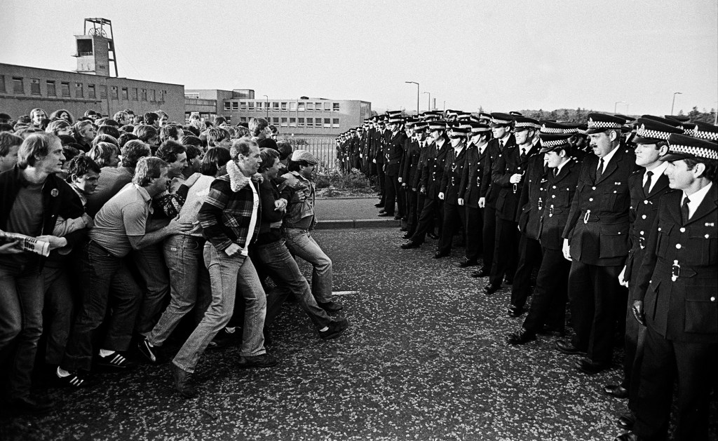 A crowd of striking miners on the left of the picture line up facing an equally large group of police men. A single miner appears to be holding back the crowd.